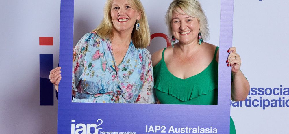 Two people smiling with a banner that reads IAP2 conference