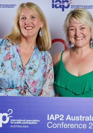 Two people smiling with a banner that reads IAP2 conference